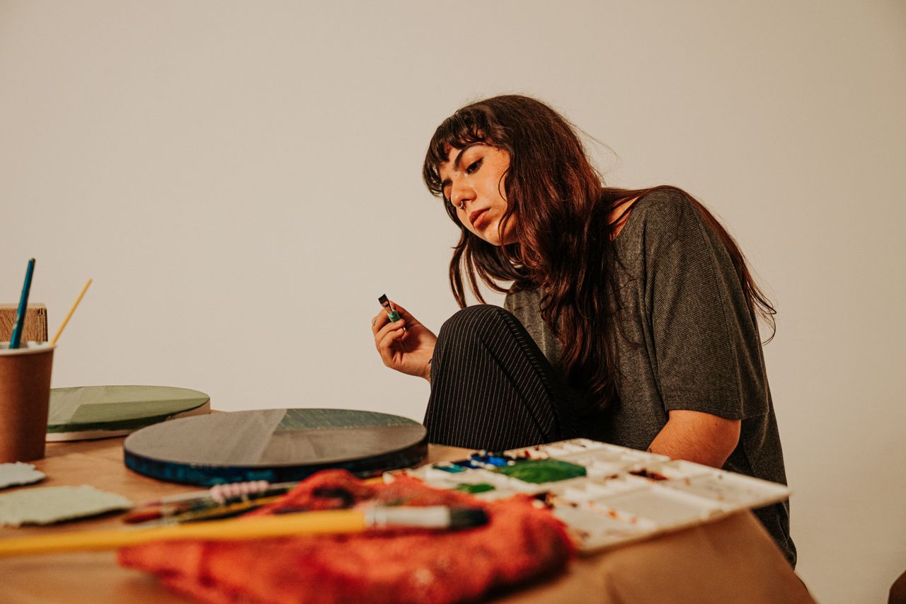 service-03 A thoughtful female artist holds a paint bottle while surrounded by her tools in an art studio.