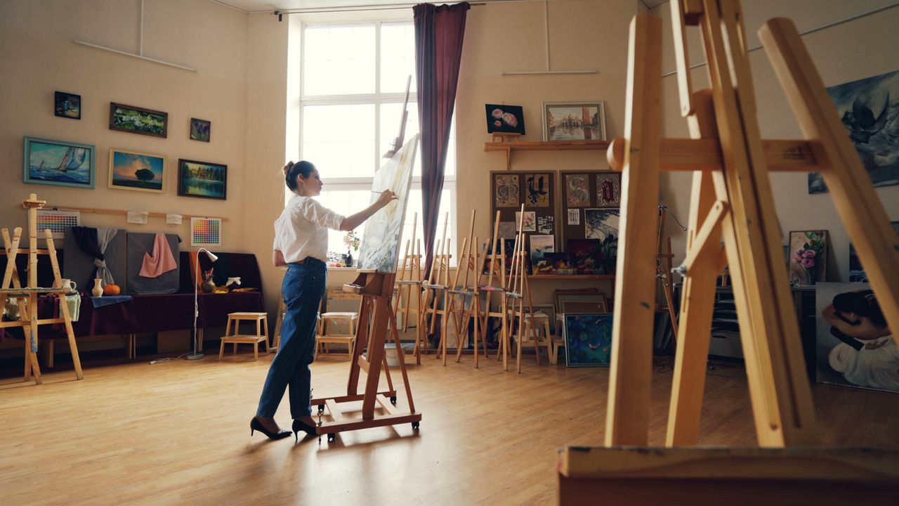 hero-img Woman painting on canvas in a sunlit art studio with easels and artwork.