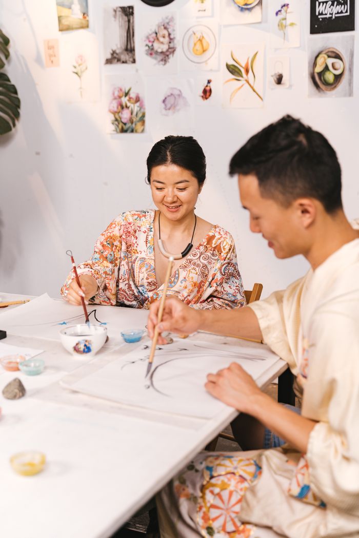 about-img Two adults engaging in a Japanese art class, painting with brushes in a bright studio.